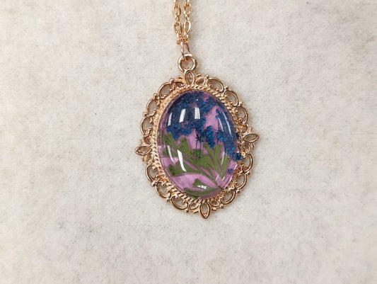 A necklace featuring an enamel floral design with a gold-colored metal chain and a central pendulum with a floral pattern made of real flowers.